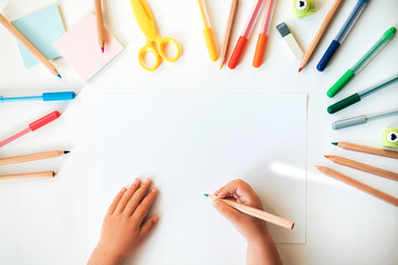 Close up of child's hands drawing at white paper within colorful pens and pencils.