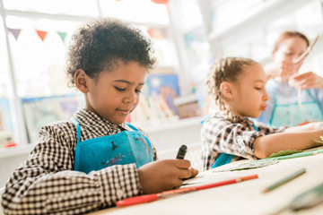 Cute African schoolboy in blue apron using highlighter while drawing at lesson