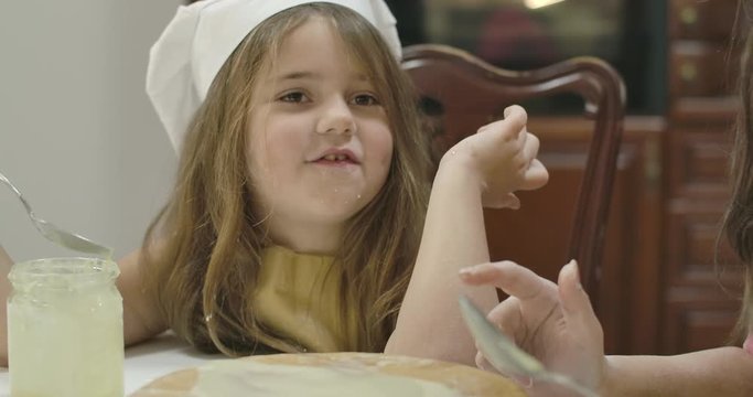 Cake smeared with condensed milk standing at table and little pretty girl eating sweetened milk at the background. Cute Caucasian daughter helping her mother at kitchen. Cinema 4k footage ProRes HQ.