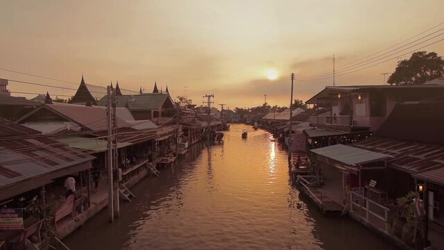 Vdo. river view evening of a long-tail boat running in canal around with Thai wooden house on both bank with yellow sun light background, sunset at Amphawa Floating Market, Samut Songkhram, Thailand.
