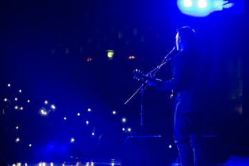 A young man with guitar sings in front of a hall full of lights and flashes