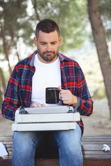 Fototapeta premium Hombre escritor con lentes escribiendo un libro a máquina de escribir. Tomando una taza de café en medio del bosque para encontrar inspiración.