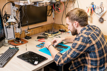 Fototapeta na wymiar Professional repairman with tweezers bending over broken smartphone by workplace