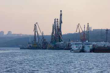 Fototapeta premium View on industrial landscape with cranes in a port. Cranes are near a river to load and unload cargo vessels.A place of a real business and industry.