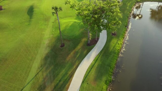 Aerial: Drone descending over empty footpath amidst trees on green golf course by pond during sunset - Kiawah Island, SC