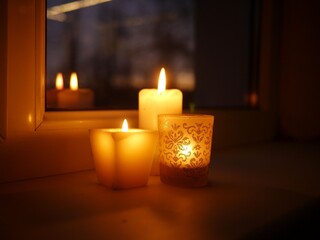 Closeup photo of three lit candles - two paraffin wax candles and one tealight in a glass candle holder with glittery golden ornament, standing on a windowsill late in the evening.