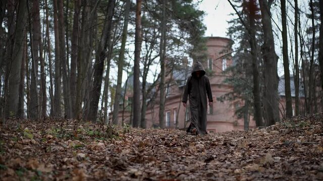 Mysterious monk or wizard in hooded robe walks throw autumn forest from medieval castle or monastery. 