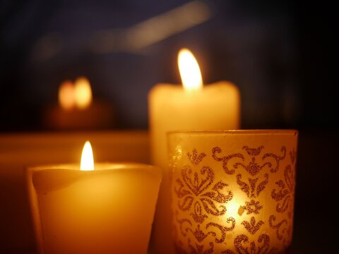 Closeup photo of three lit candles - two paraffin wax candles and one tealight in a glass candle holder with glittery golden ornament, standing on a windowsill late in the evening.