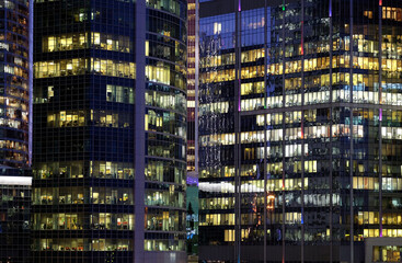 Fototapeta na wymiar Glass walls of modern office buildings with many large panoramic windows in business cluster with working people front view at night