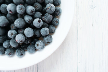 White plate with frozen chokeberry berries on a wooden white table. Top view. Copy space.