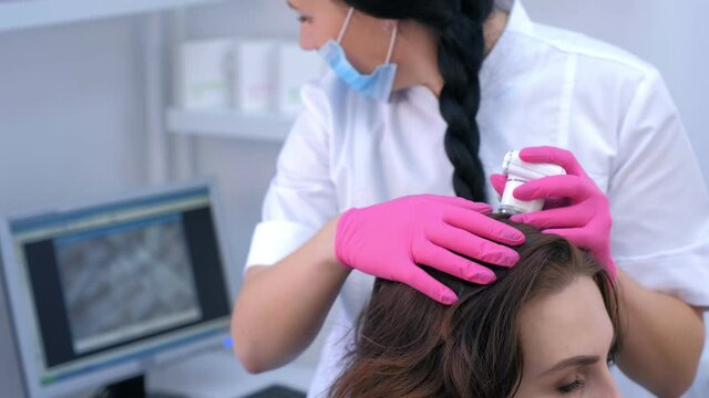 Doctor trichologist examines woman patient's hairs using trichoscope in clinic. Procedure of trichoscopy hair. Runs a trichoscope over scalp and looks result on monitor screen. Computer trichoscopy.