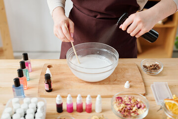 Hands of female spraying aromatic oil into liquid soap mass in glassware