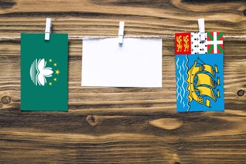 Hanging flags of Macao and Saint Pierre And Miquelon attached to rope with clothes pins with copy space on white note paper on wooden background.Diplomatic relations between countries.