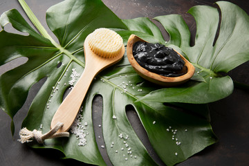 Spa. Dead sea mud in a bowl of olive wood, brush for body massage, sea salt on big green leaf. On dark background top view. Copy space
