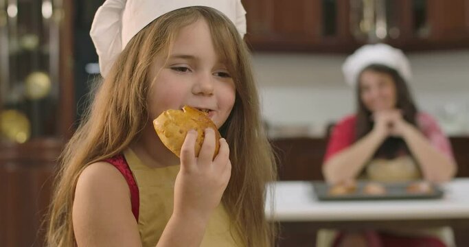 Little pretty Caucasian girl eating bun at the kitchen at home. Cute child enjoying tasty bakery as her mother sitting at the background and smiling. Cinema 4k footage ProRes HQ.