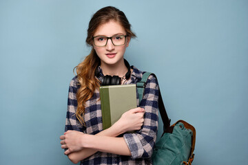 A girl in a plaid shirt and glasses with headphones on neck stands with a backpack, clutching a book to herself.