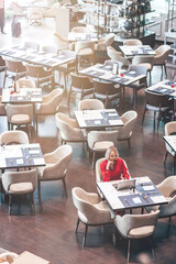 Portrait of gorgeous lady with blonde hair wearing red jumpsuit, looking at the screen of her laptop while sitting in an empty restaurant. Successful beautiful woman concept. Vertical shot. Top view