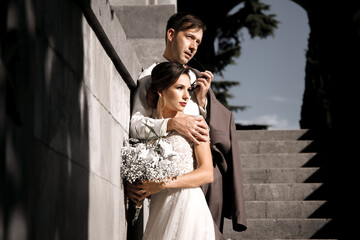 Happy groom look to beautiful bride wearing wedding dress and holding marriage bouquet in old street of the city with beautiful view. The couple hugging in love.