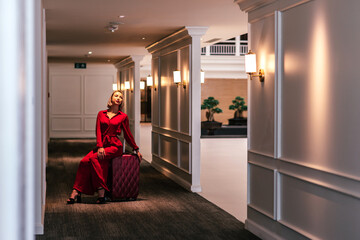 Full-length portrait of gorgeous lady with blonde hair wearing red jumpsuit sitting on her suitcase while waiting in the hall of modern hotel. Business trip concept. Horizontal shot