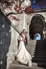 Obraz premium Happy groom look to beautiful bride wearing wedding dress and holding marriage bouquet in old street of the city with beautiful view. The couple hugging in love.