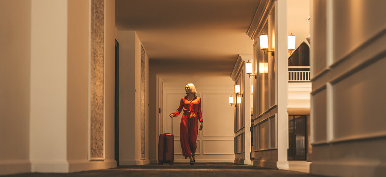 Full-length portrait of gorgeous lady with blonde hair wearing red jumpsuit walking along the hall in hotel with a suitcase. Business trip concept. Horizontal shot. Front view