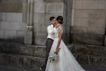 Happy groom look to beautiful bride wearing elegant wedding dress and holding marriage bouquet in old street of the city with beautiful view. The couple n love.