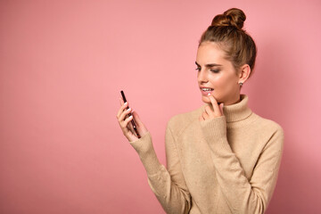 Obraz premium A girl in a beige turtleneck with gathered hair stands on a pink background and looks thoughtfully into a smartphone.