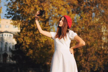 Outdoor shot of lovely girl wearing beret in autumn park makes self portrait on her cellphone. Girl holding smartphone making photo in open air for social networking. People concept