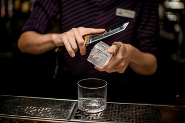 Professional bartender cutting ice with a knife above the empty cocktail glass