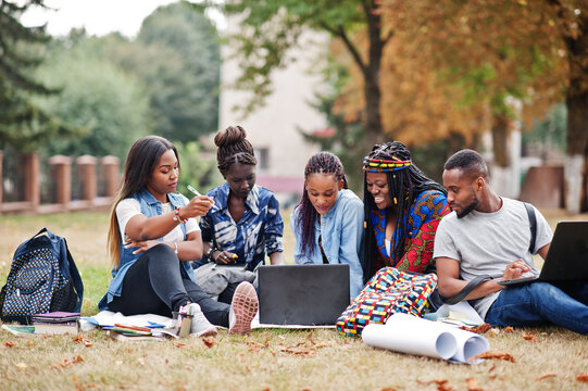 Group of five african college students spending time together on campus at university yard. Black afro friends sitting on grass and studying with laptops.