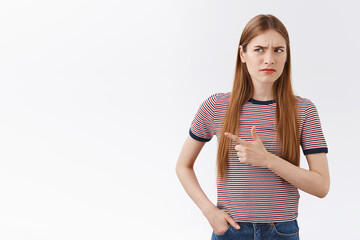 Hesitant and suspicious, unsure young fair-haired woman in striped t-shirt, frowning, squinting uncertain, pointing left and peek skeptical, standing confused white background, see something strange