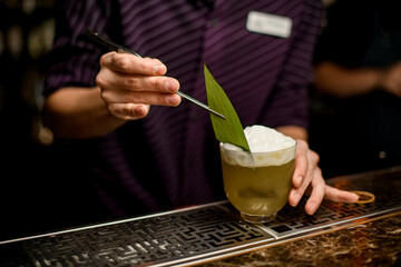 Bartender decorating a delicious yellow alcoholic cocktail in the glass with a whipped cream with a tropical leaf