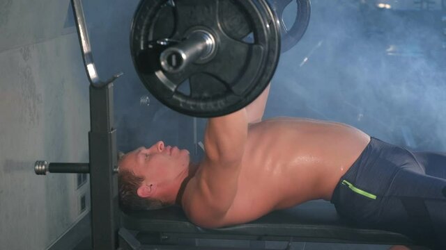 Professional muscular athletic man bodybuilder doing barbell chest exercise on bench in gym, side view. Man pumping chest with naked torso. Perfect bench press technique. White smoke in gym in dark.