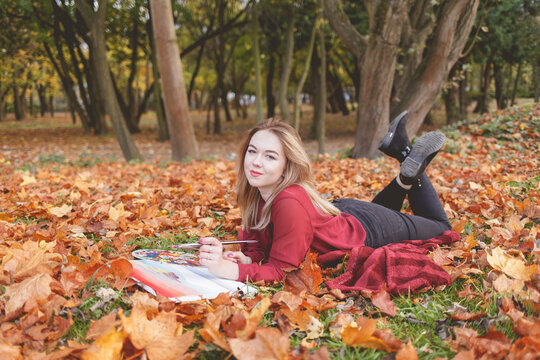 Young artist lies in autumn park on a plaid and paints a picture.  Girl draws a picture in the park