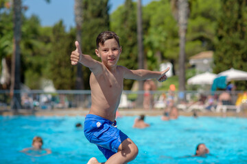 Cute active European boy in blue swimming shorts having fun in hotel&rsquo;s swimming pool, he is making fantastic jump, smiling and showing like gestures.