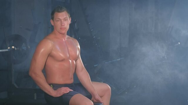 Portrait of professional muscular athletic man bodybuilder in shorts is sitting on bench in gym and looking at camera. Perfect body with naked torso. White smoke in gym in dark. Sport motivation.