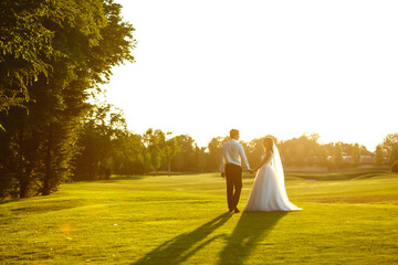 Fototapeta premium Happy bride and groom go into the sunset. Lovely wedding couple at sunset. Bride and groom in wedding attire is in the hands against the backdrop of the green field at sunset. Together. Wedding. 