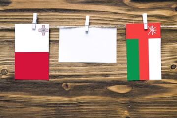 Hanging flags of Malta and Oman attached to rope with clothes pins with copy space on white note paper on wooden background.Diplomatic relations between countries.