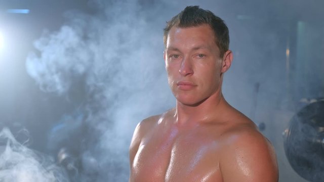 Portrait of professional muscular athletic man bodybuilder is sitting on bench in gym and looking at camera. Perfect body with naked torso. White smoke in gym in dark. Sport motivation concept.