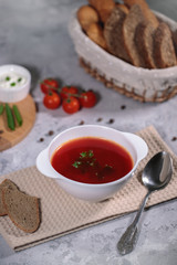 Tasty and hearty dinner. A plate with borsch on the table, next to the board are vegetables, parsley, dill, green onions, garlic, chili peppers, cherry tomatoes, and bread.