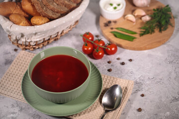 Tasty and hearty dinner. A plate with borsch on the table, next to the board are vegetables, parsley, dill, green onions, garlic, chili peppers, cherry tomatoes, and bread.