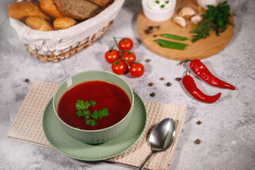 Tasty and hearty dinner. A plate with borsch on the table, next to the board are vegetables, parsley, dill, green onions, garlic, chili peppers, cherry tomatoes, and bread.