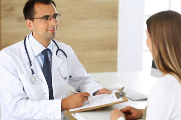 Doctor and patient discussing the results of a physical examination while sitting at a desk in a clinic. A male doctor using a clipboard to fill out a medical history of a young woman's medication