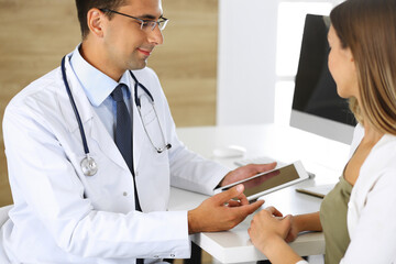 Doctor and patient discussing medical exam results while sitting at the desk in clinic. Male physician using tablet computer for filling up medication history records of young woman. Data in medicine