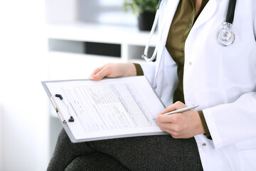 Woman doctor writing something at clipboard while sitting at the chair, close-up. Therapist at work filling up medication history records. Medicine and healthcare concept