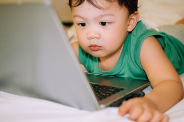 A male toddler is playing with a laptop while on the bed