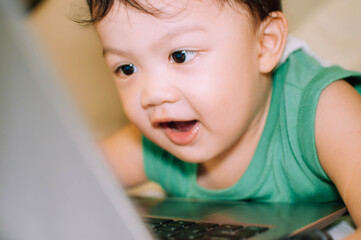 A male toddler is playing with a laptop while on the bed