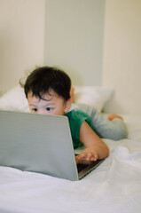 A male toddler is playing with a laptop while on the bed