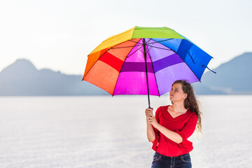 Young happy woman holding colorful rainbow umbrella for shade on white Bonneville Salt Flats near Salt Lake City, Utah and mountain view