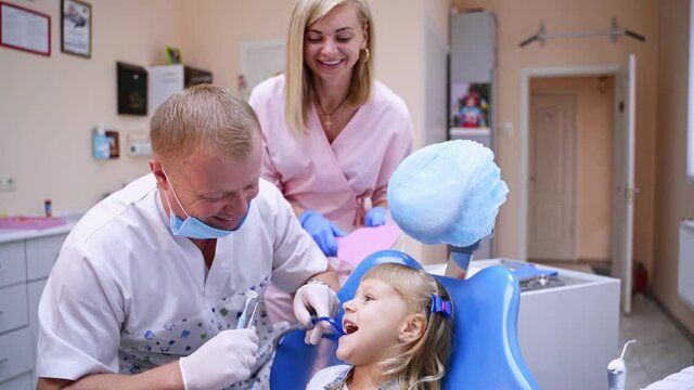 Smiling girl at dentist's visit. Little child is sitting in dentist chair. Doctor treats girls teeth with his assistant on the modern medical room background.